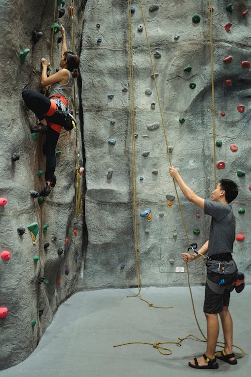 Side view of anonymous young ethnic male trainer in activewear holding belay rope while female climber ascending wall in gym