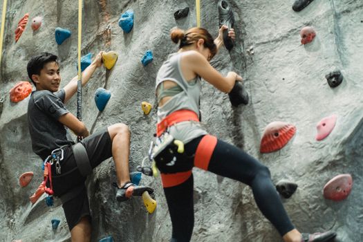 Low angle of young Asian male and female friends in sportswear and protective harness practicing bouldering together in climbing gym