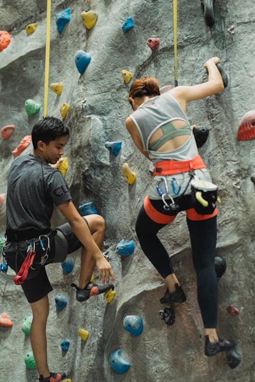 Free From below back view of young Asian male mountaineer with unrecognizable sportswoman climbing wall with grips while practicing extreme sport Stock Photo