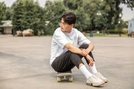 Full body of pensive ethnic male teenager in trendy outfit and eyeglasses sitting o skateboard and looking away while resting in park after training