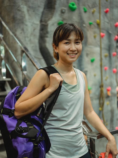 Smiling Asian climber with sports bag near artificial rock