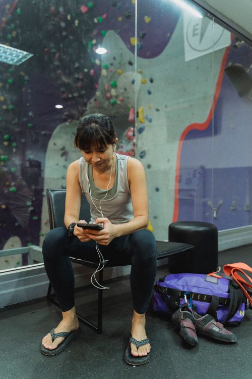 Free Young smiling ethnic female mountaineer in earphones text messaging on cellphone near professional equipment and climbing wall Stock Photo