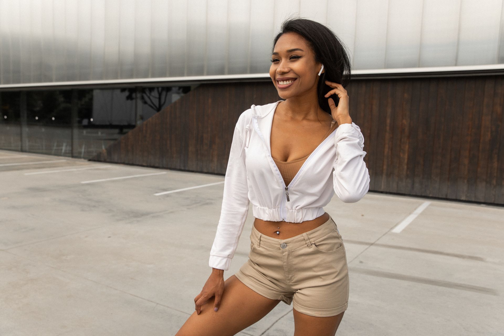 Confident fit young ethnic woman in trendy outfit touching long dark hair while listening to music in true wireless earphones standing near modern building on street