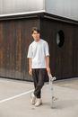 Full length of confident young Asian guy in white t shirt and eyeglasses leaning on skateboard while standing in city park after workout