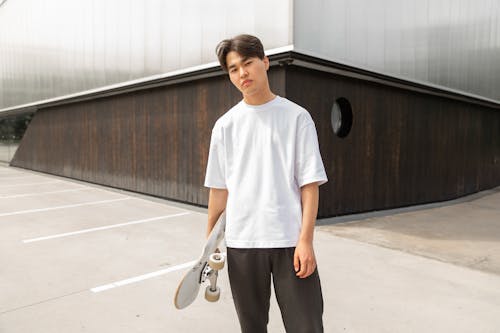 Confident young Asian man standing near modern building with skateboard in hand