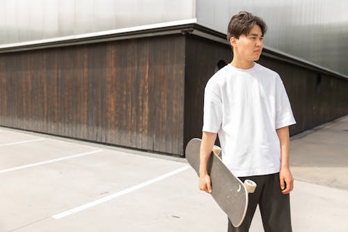 Handsome young ethnic guy in white t shirt holding skateboard in hand and looking away thoughtfully while resting near modern building after ride