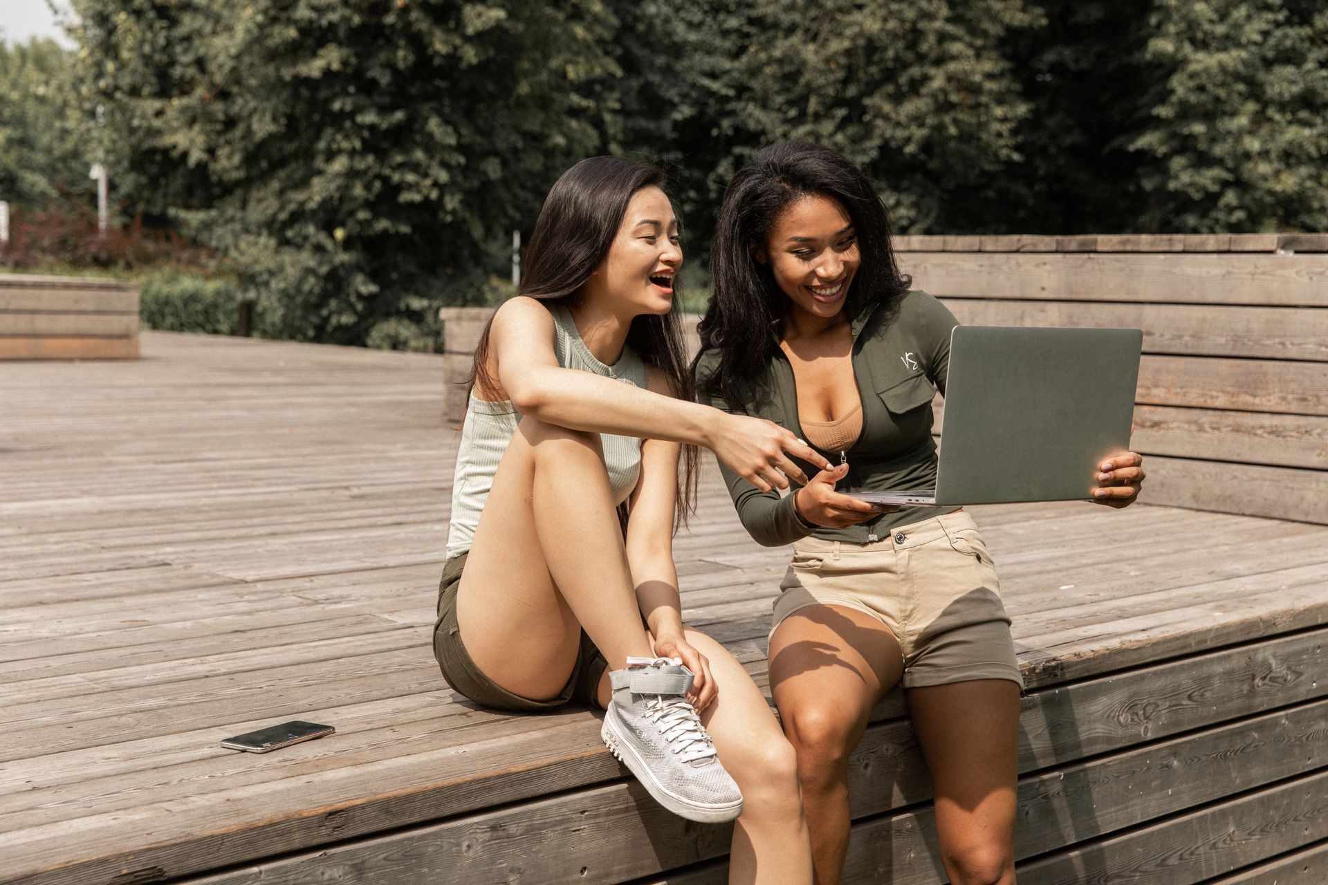 Happy young multiracial female friends in casual clothes laughing while watching funny video on laptop sitting on wooden podium in park