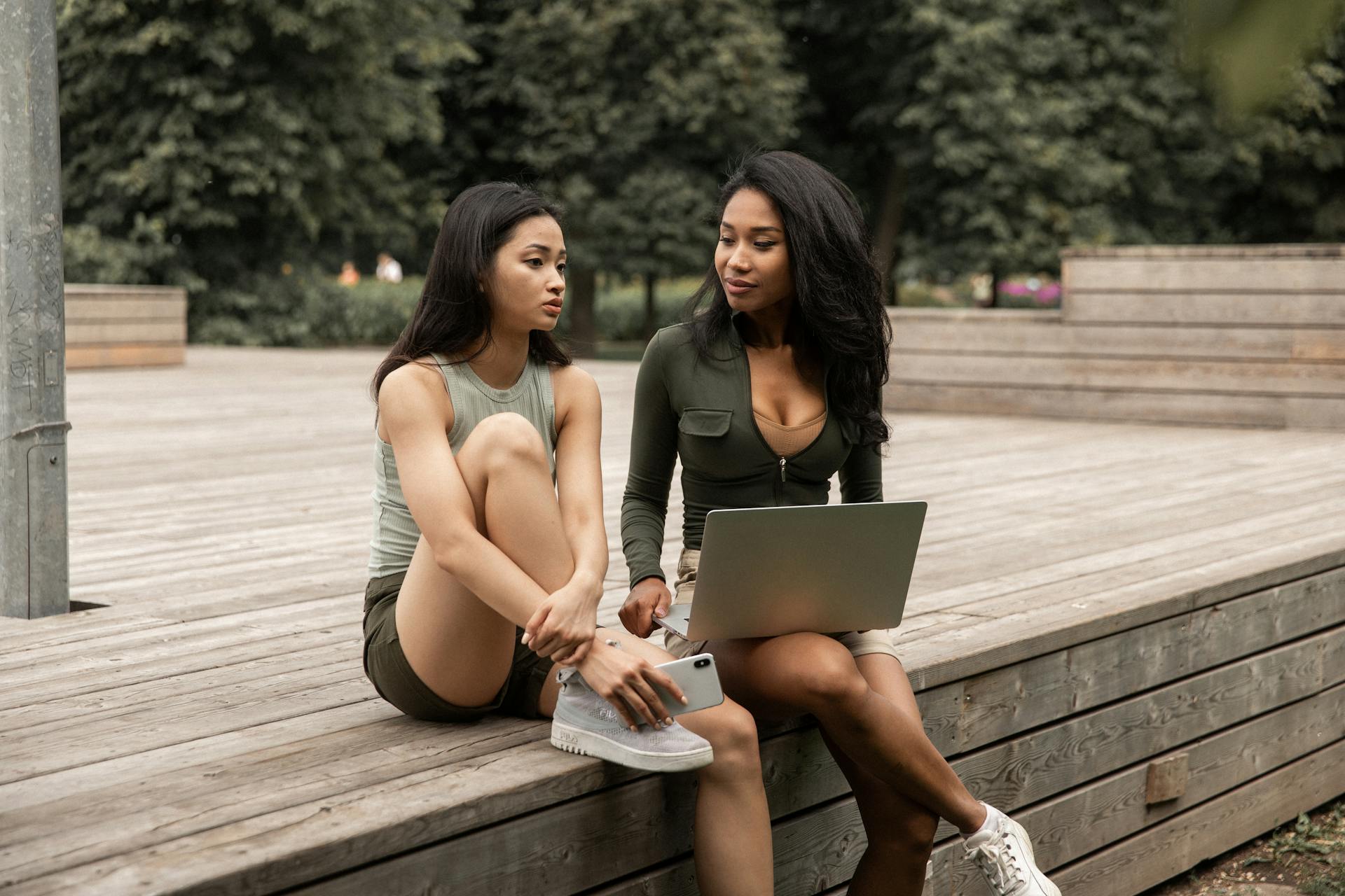 Two women enjoying a casual conversation outdoors, sharing a laptop and smartphone.