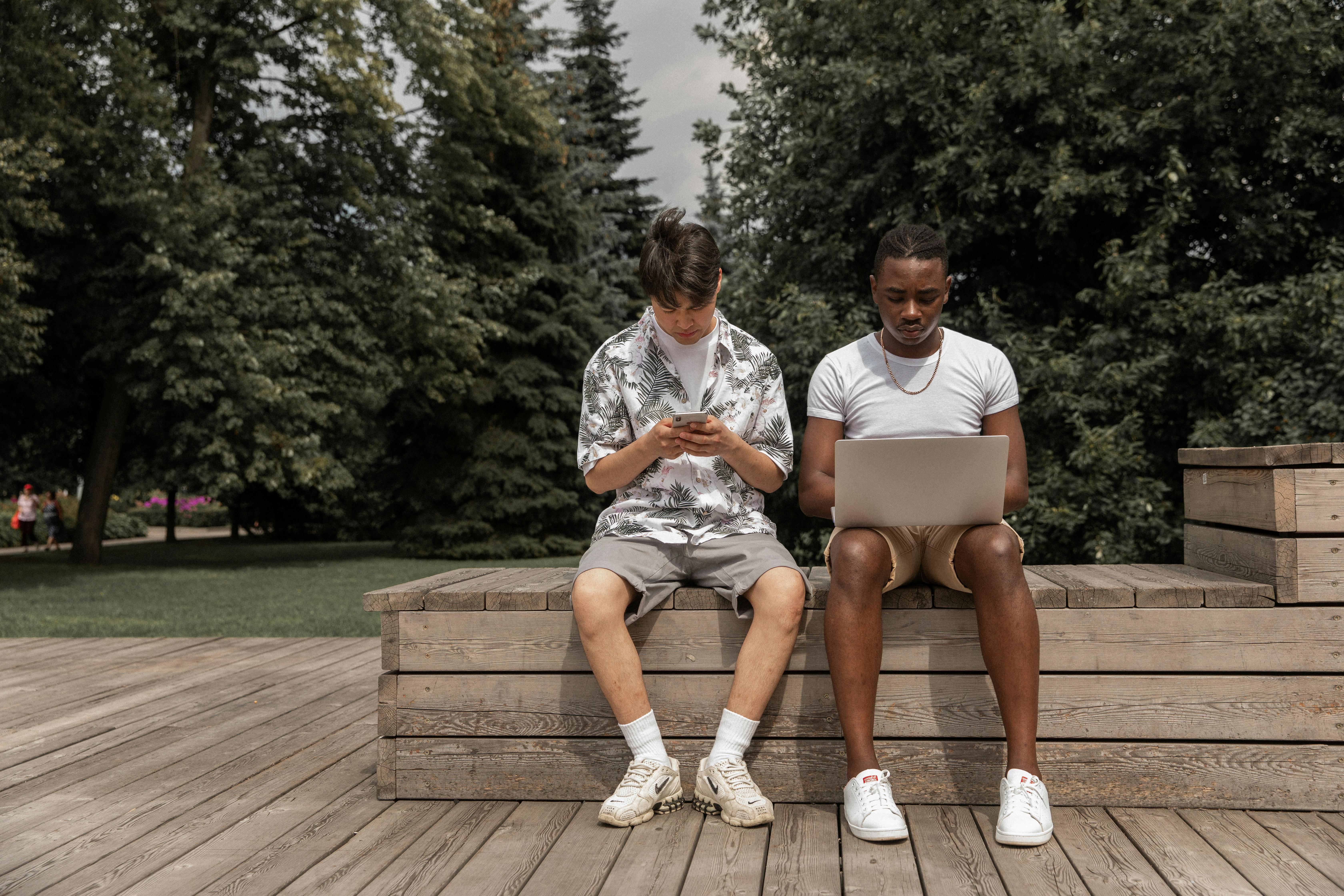 concentrated young diverse men using smartphone and netbook sitting on bench in park