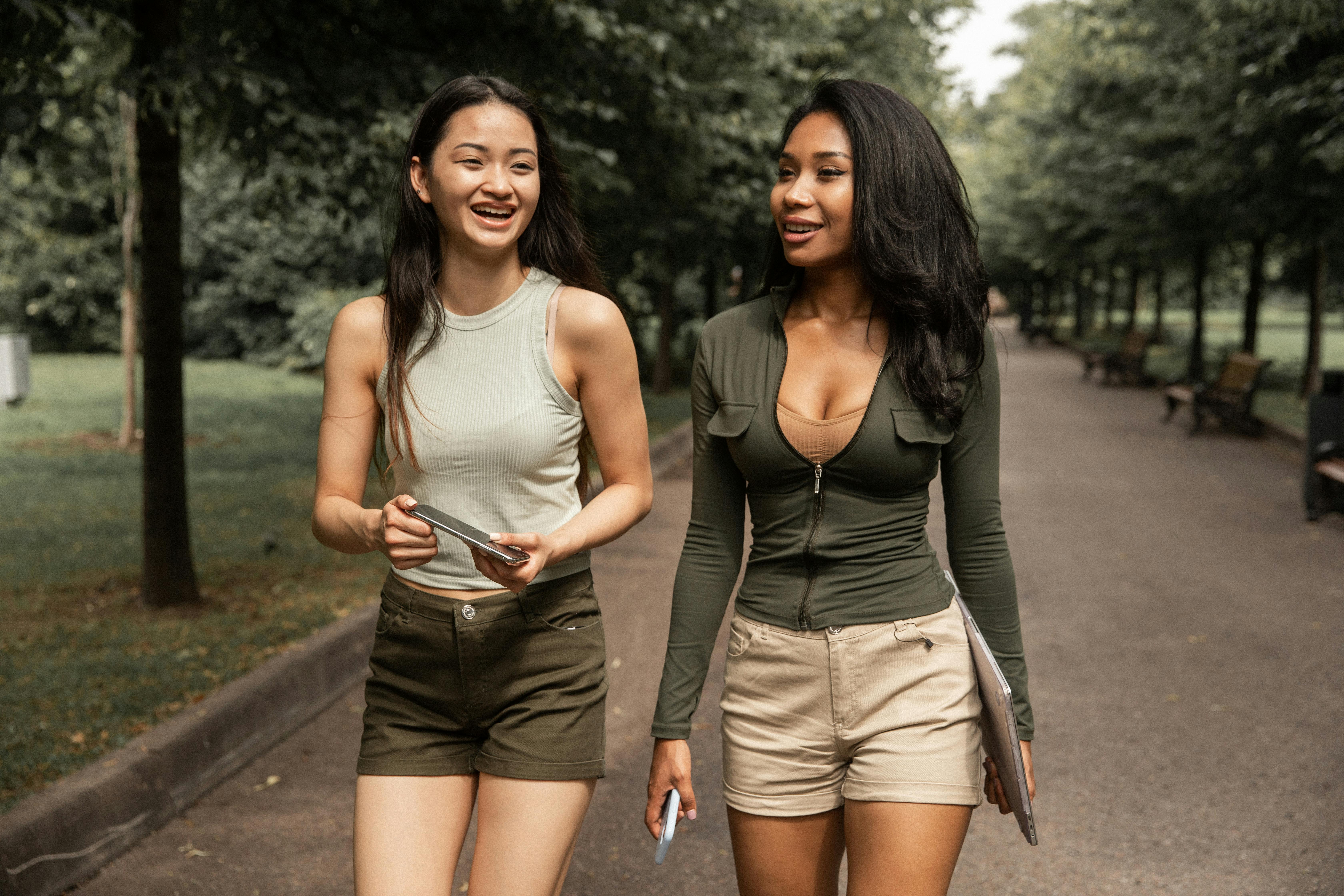 positive young diverse women walking along path in park with gadgets in hands