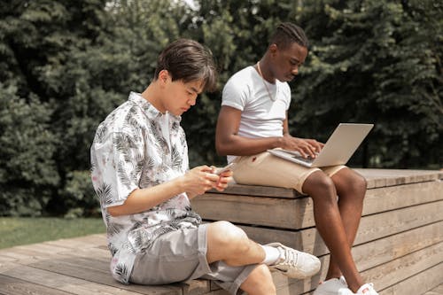 Side view of focused young multiracial male friends sitting on wooden bench and working remotely in park using laptop and smartphone on sunny day