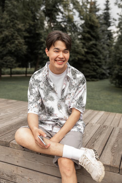 High angle of positive young ethnic man in trendy outfit smiling and looking at camera while relaxing on wooden bench in green park