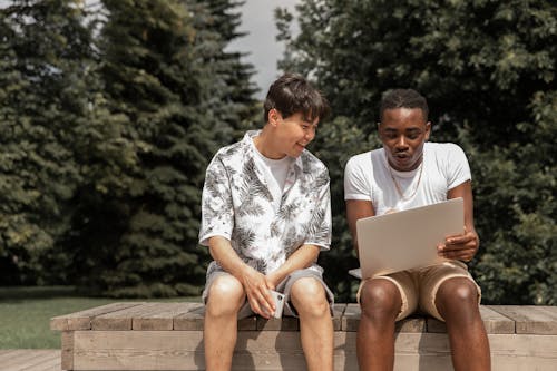 Smiling multiracial friends in stylish clothes sitting on wooden bench and discussing information from netbook