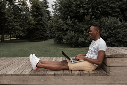 Side view of African American male in casual outfit sitting on wooden surface and browsing netbook while looking at screen