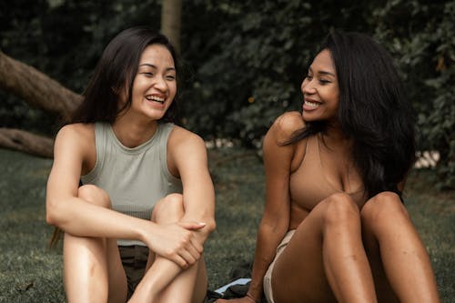 Free Charismatic multiracial girlfriends in summer clothes sitting on grass and laughing while having meeting in park Stock Photo