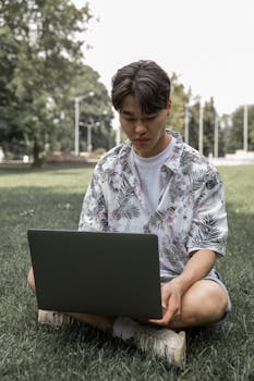 A young man sits cross-legged on grass, focused on a laptop in a sunny park.
