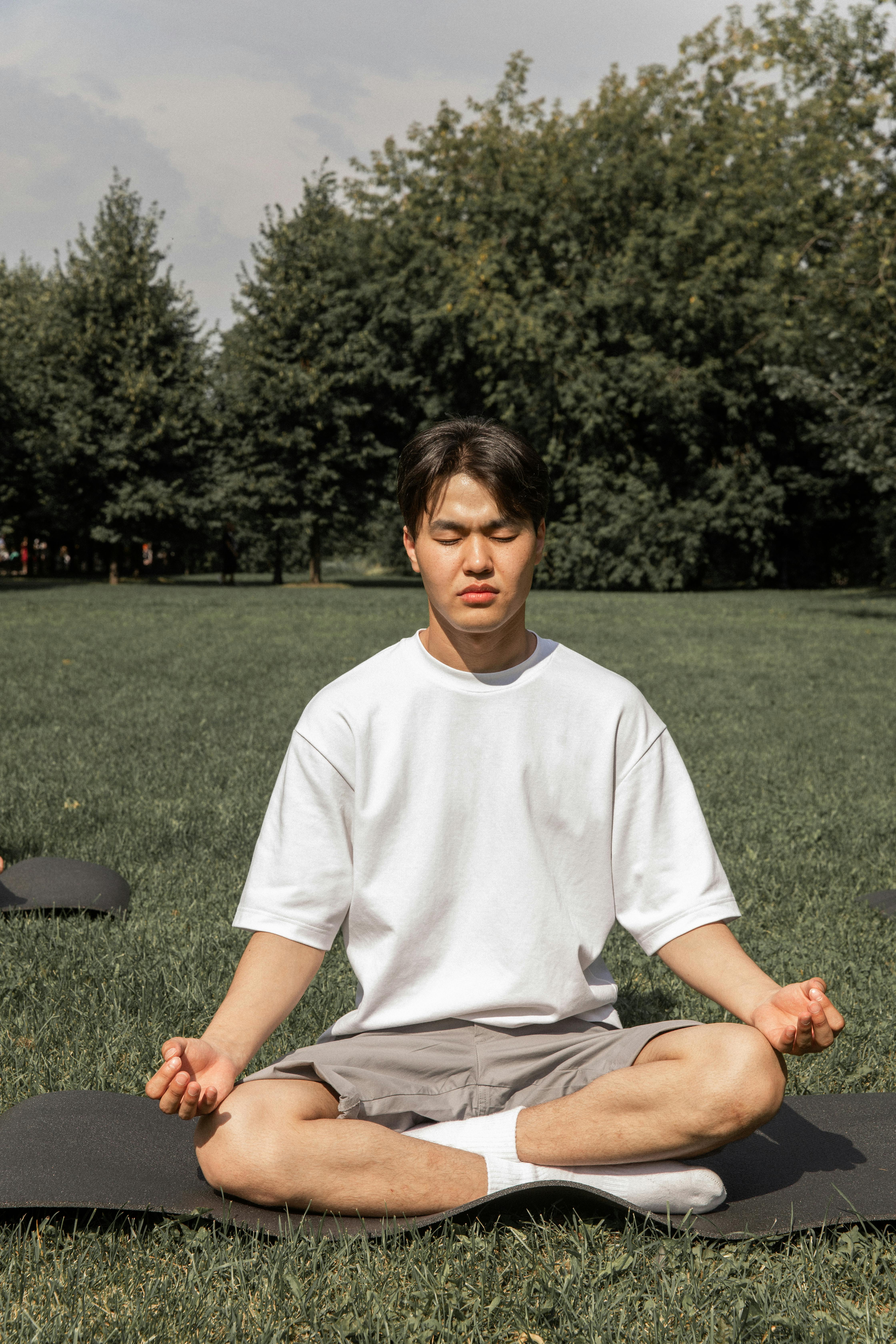 Full body of concentrated Asian male in casual wear sitting in Padmasana with closed eyes in summer park
