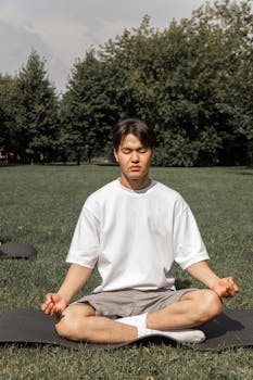 Full body of concentrated Asian male in casual wear sitting in Padmasana with closed eyes in summer park