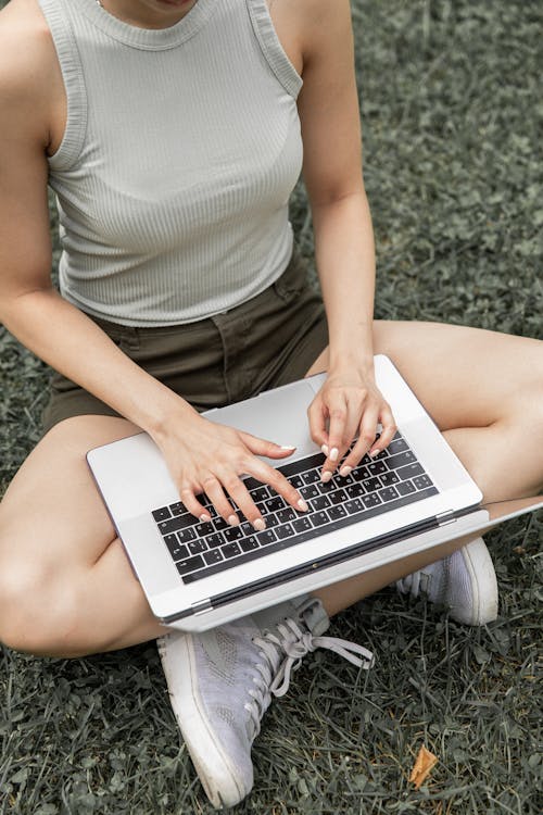High angle of unrecognizable female in casual outfit sitting on lawn with crossed legs and studying online