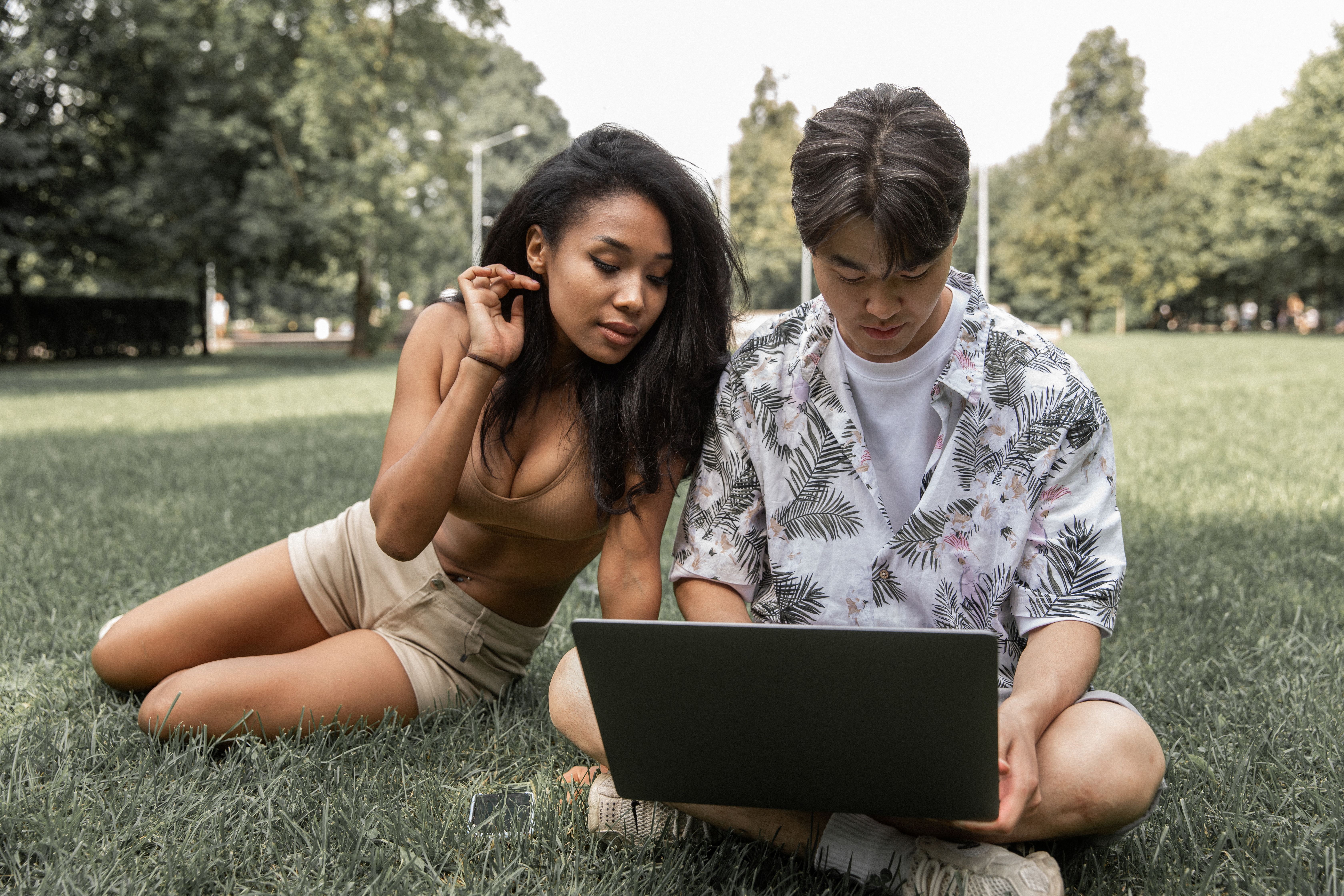 Full body of multiethnic couple in summer clothes relaxing on grassy lawn while browsing netbook together and looking at screen
