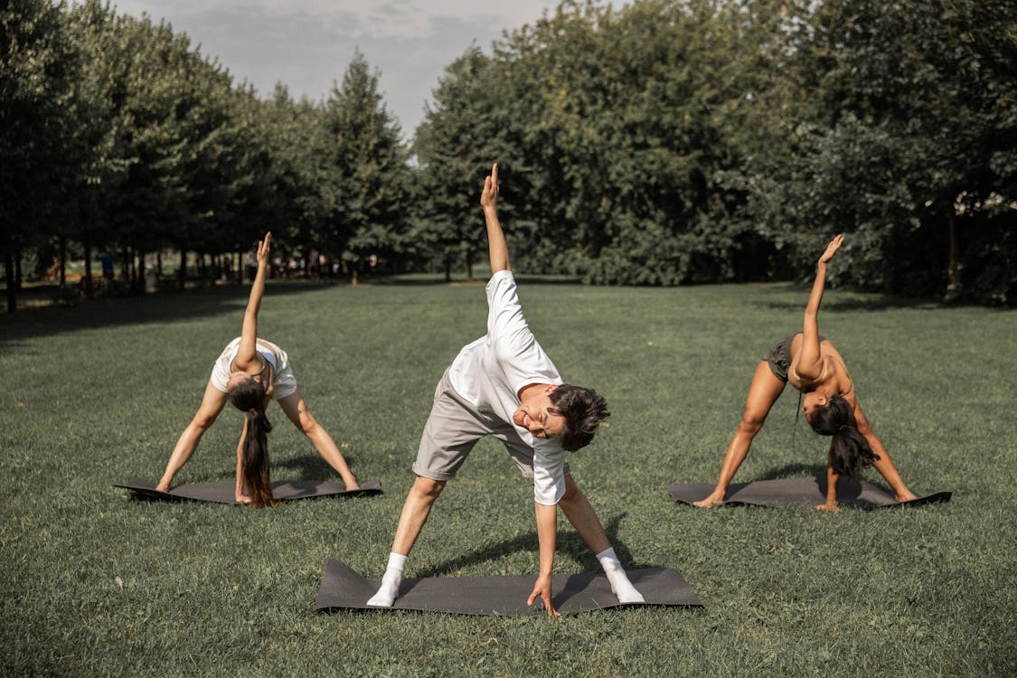 Group of diverse friends doing Revolved Wide Legged Forward Bend while exercising yoga on lawn