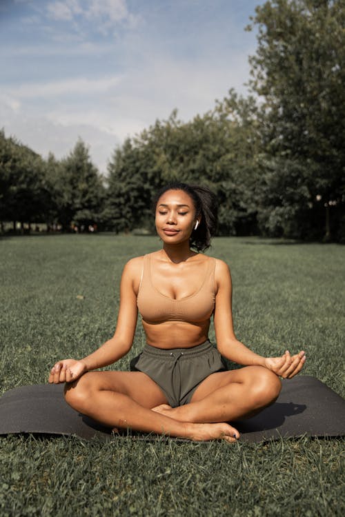 Free Full length of barefoot ethnic female in activewear sitting in lotus pose with closed eyes while listening to music in earbuds and meditating Stock Photo