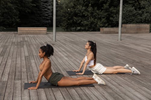 Free Side view of focused diverse girlfriends doing High Cobra pose while stretching bodies on wooden platform in park Stock Photo
