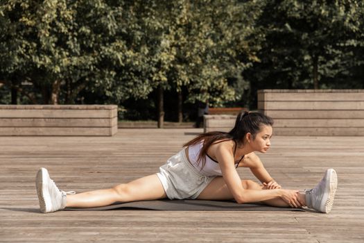 Full body of Asian female in activewear stretching legs while practicing yoga on wooden platform in garden