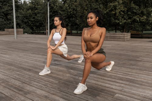 Free Full body of focused multiracial girlfriends doing Crescent Lunge with hands on knee on wooden podium in summer garden Stock Photo