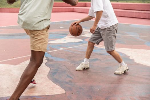 Crop anonymous multiethnic fit men in sportswear playing basketball on sports ground on clear summer day