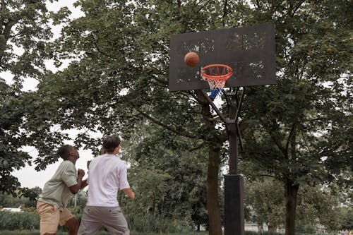Unrecognizable diverse sportsmen in activewear throwing ball in hoop while playing basketball on sports ground in summer park
