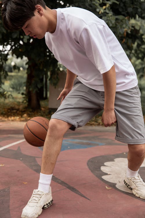 Crop focused Asian sportsman in activewear dribbling ball while playing basketball on sports ground on clear summer day