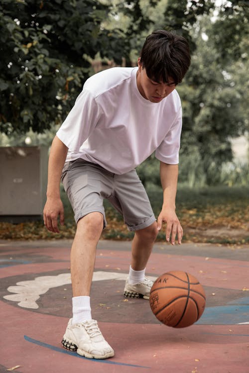 Full length sporty ethnic sportsman in light activewear dribbling basketball ball on sports ground in green city park