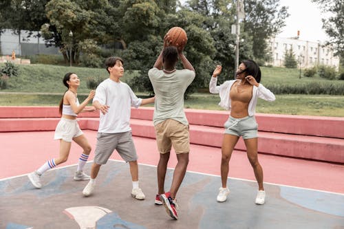 Full length sporty young friends wearing activewear playing basketball together on sports ground in green city park