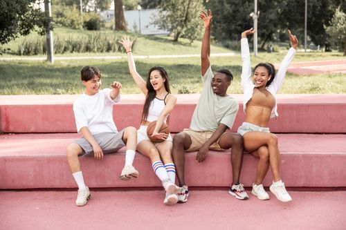 Free Full body joyful young diverse friends sitting on pink stone bench in lush sunny park and raising arms in excitement Stock Photo