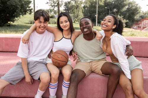 Free Content young multiracial friends wearing casual clothes embracing and looking at camera with happy smiles while sitting on bench in green park Stock Photo