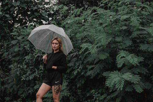 Young focused female in trendy apparel standing with transparent umbrella near greenery plants in garden while looking at camera