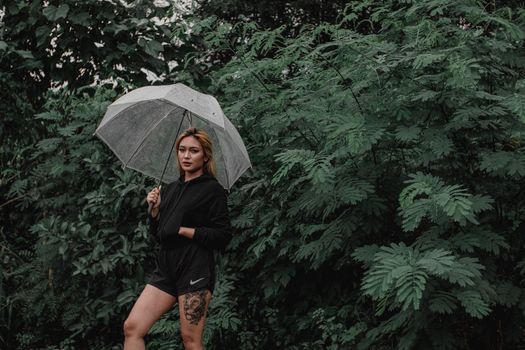 一位穿着时尚的年轻女性专注地站在花园的绿叶植物旁，举着透明伞看向镜头