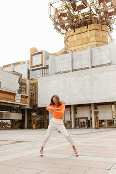 一位身着鲜艳橙色和白色衣服的女子，在现代城市建筑前微笑着跳舞