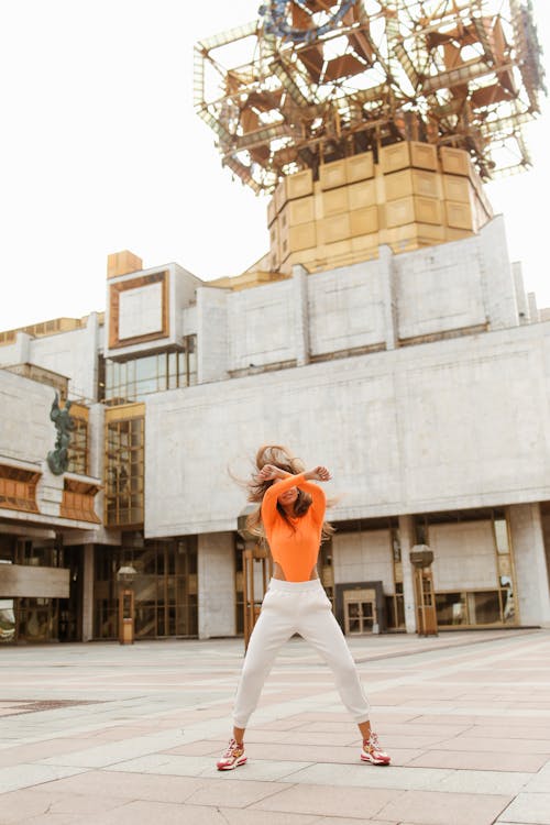 Free Woman in Orange Long Sleeves and White Pants Dancing Cheerfully  Stock Photo