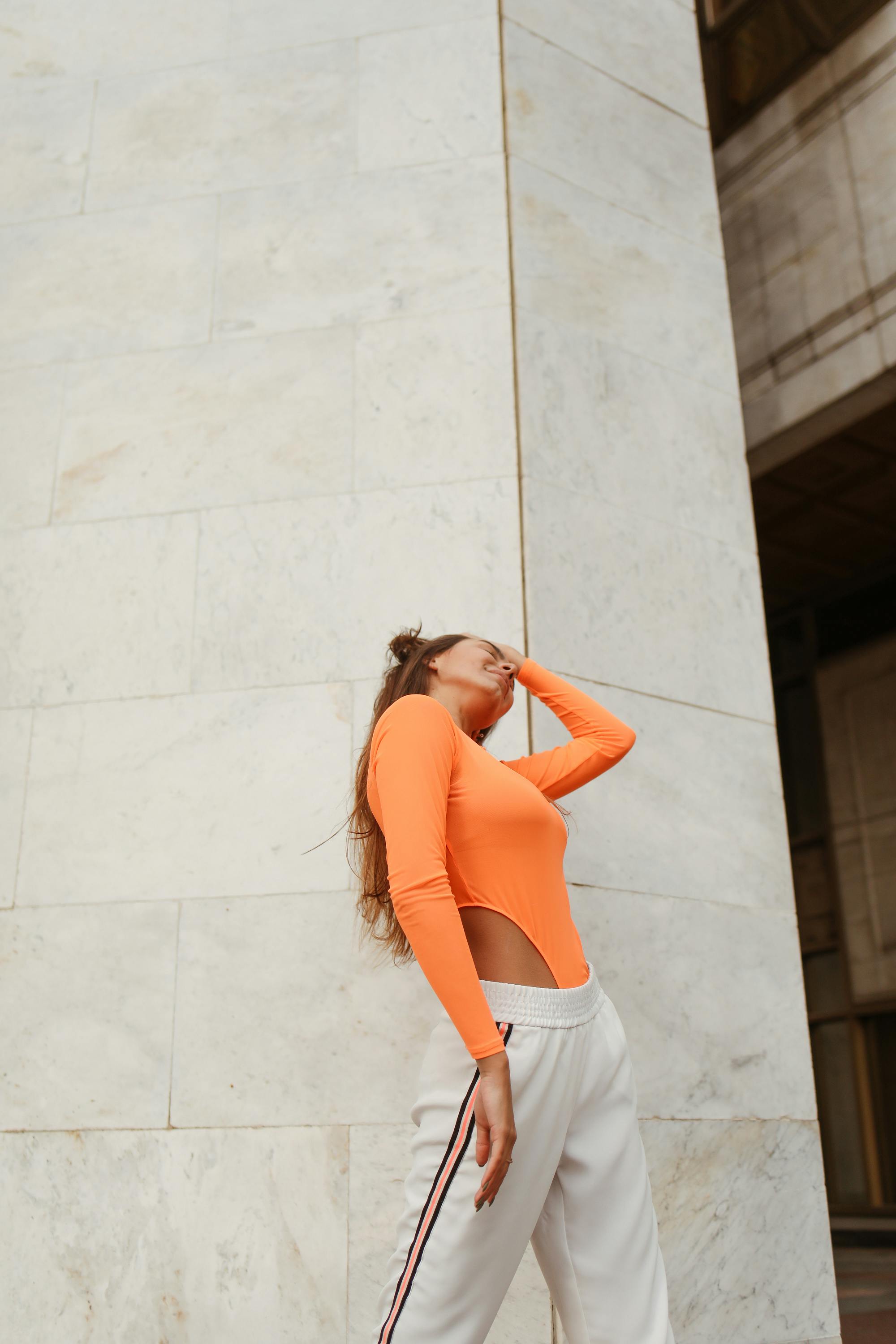 一位身着橙色紧身衣和白色裤子的时髦女性，倚靠在户外的大理石墙边
