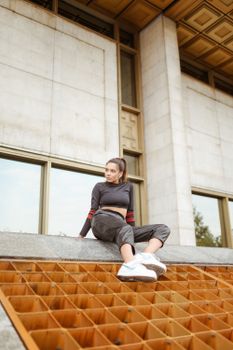 一位穿着休闲装的时尚女士坐在现代建筑外的结构上，展现街头服饰的潮流