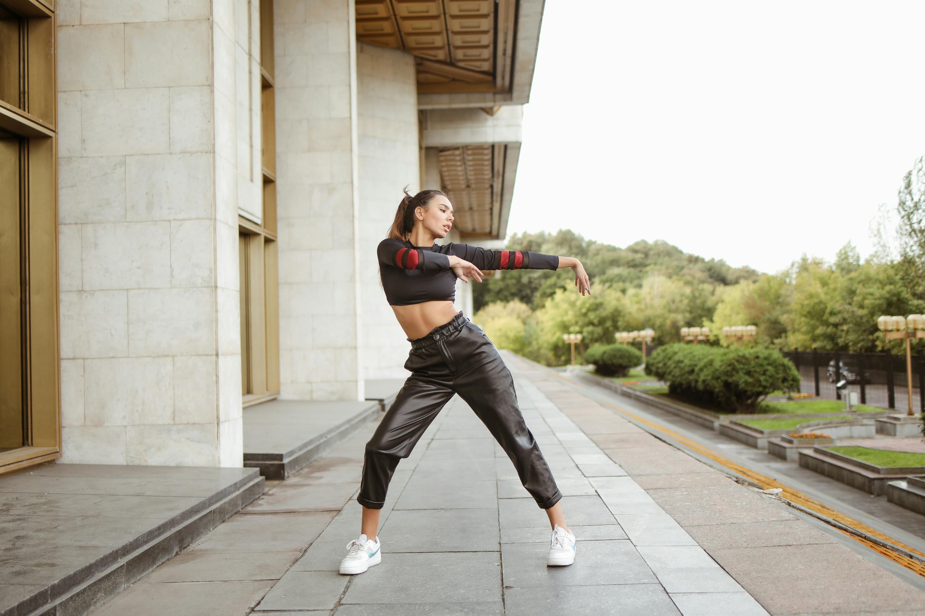 Female dancer in black streetwear poses energetically in urban outdoor setting.