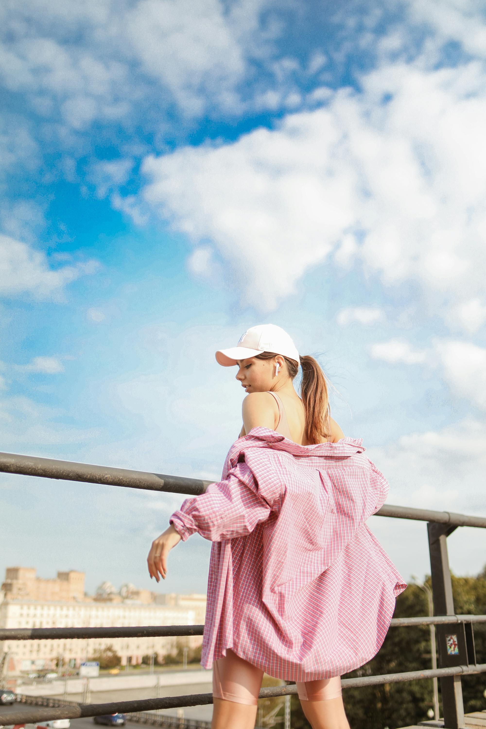 一位时尚女性穿着粉色超大衬衫、戴着帽子，在明亮的天空下于户外摆姿势
