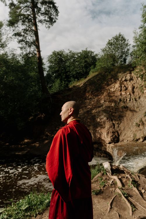 Free Bald monk in red robe stands peacefully by a tranquil river surrounded by lush trees. Stock Photo