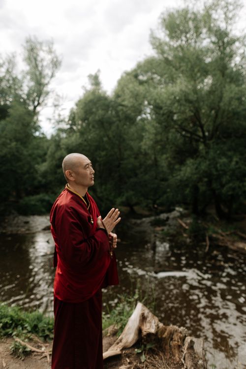 Free A monk in red robes standing in prayer by a calm river surrounded by lush greenery. Stock Photo