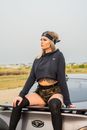 Young contemplative tattooed female in trendy apparel sitting on contemporary car while looking away in countryside