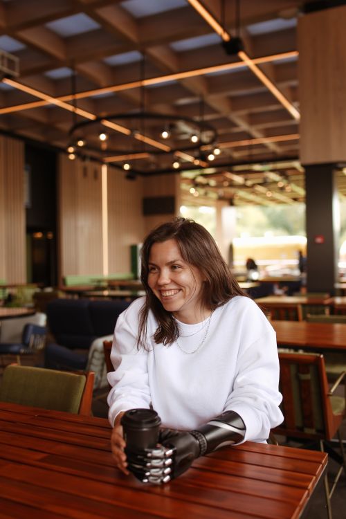 Free Young woman with prosthetic arm smiles while sitting in a cafe holding a coffee cup. Stock Photo