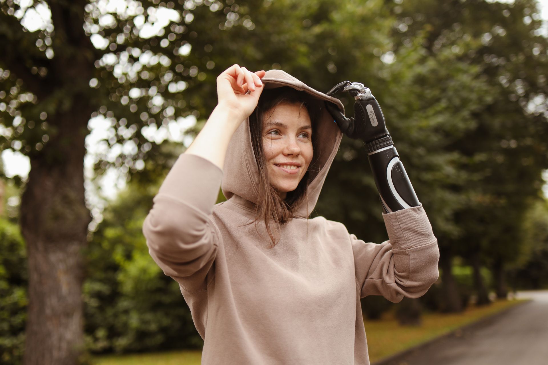 A woman with a prosthetic arm adjusts her hoodie outdoors in a serene park.