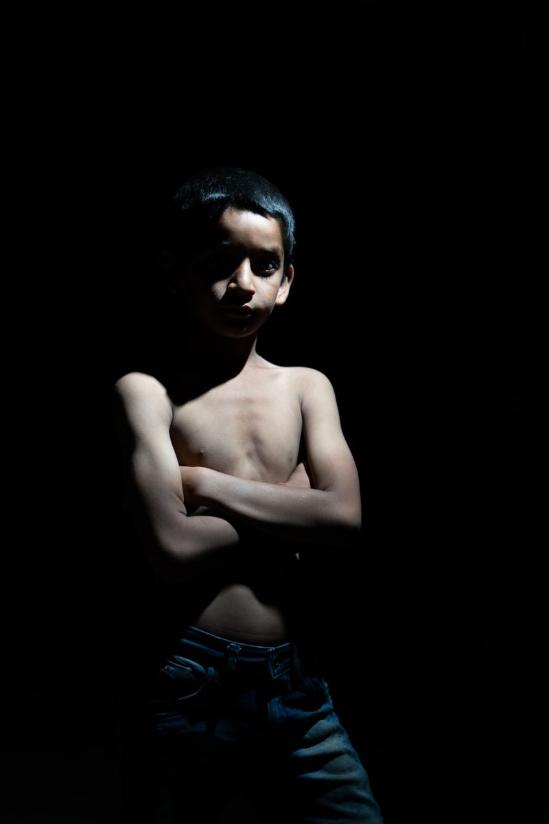 Dramatic portrait of a boy standing with arms crossed in dark lighting, creating a powerful silhouette.